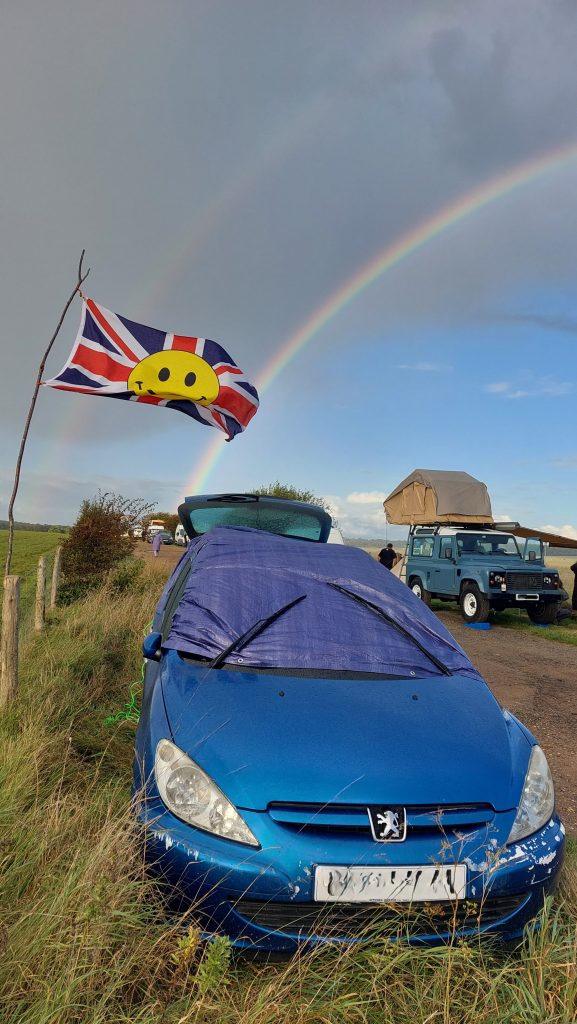 The blue tarp-covered car is facing the camera and other camping vehicles can be seen on the other side of the bridleway. The wally flag still flies, and a rainbow, with a feint double arc, beams across the sky