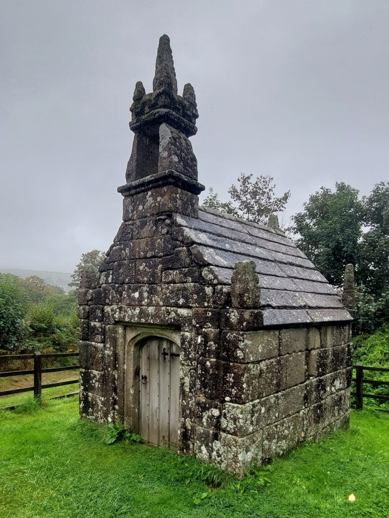 Taken on a rainy day, this view of Dupath Well Chapel shows the front of the granite building with it's 'sentinal-like spire' and one side of the roof and wall. It is set on closely cropped grass, with a wooden fence and trees in the backgroud.