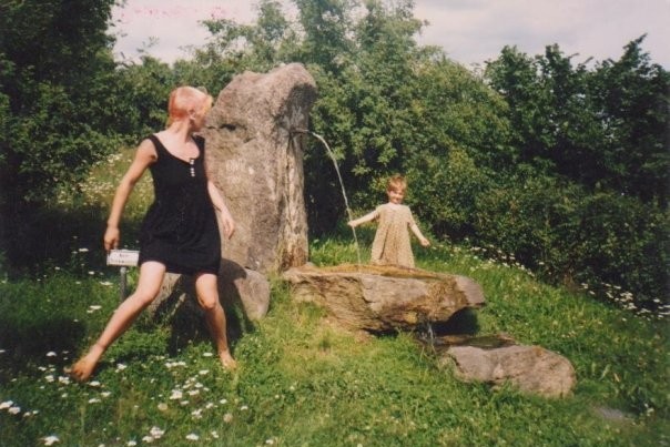 A young woman (the author) is caught mid-turn, looking back at her 5 year old daughter who stands with arms outstretched beside a rocky spring. They are surrounded by grass, wildflowers and trees