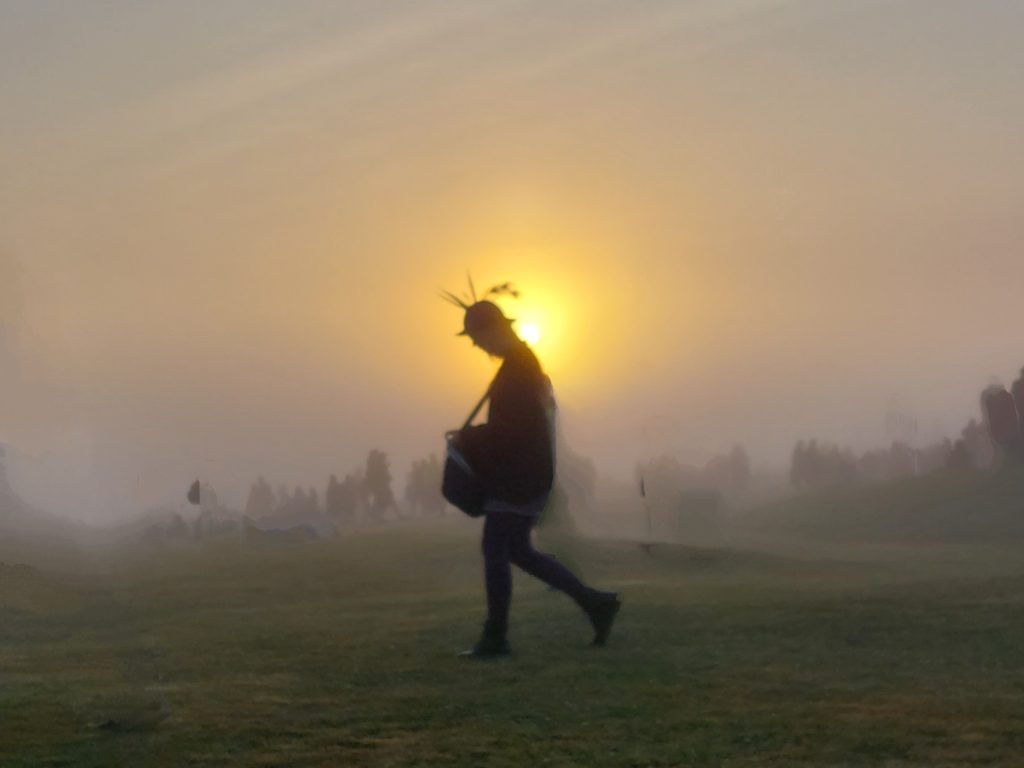 The silhouette of a lone figure, with feathered cap, walks across the grass in front of the rising sun. In the misty backgroud, there are other figures standing around in groups.