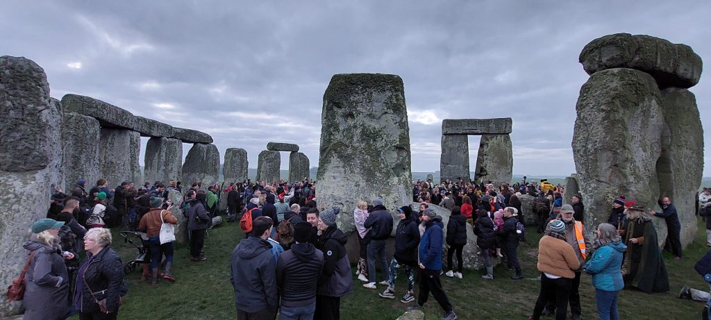 A crowd of people gathered amonst the monoliths at Stonehenge. The sky is overcast and grey. March 2024