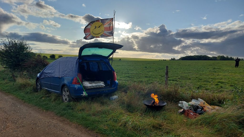 A blue estate car, covered by a blue tarp, sits on the verge of a bridleway with the hatchback open, revealing a makeshift bed inside. The 'wally flag' (a union jack with an acid-house smiley face in the middle) flaps in the wind, while a small fire burns in a fire pit