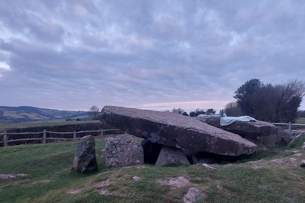 A cloudy dawn breaks across the sky above Arthur,s Stone, a collapsed burial chamber dating from around 3700-2700BC. The huge capstone sits slightly askew on top of supporting upright stones. There is a blanket and flasks on top of the capstone, where the author/photographer has been sitting :)