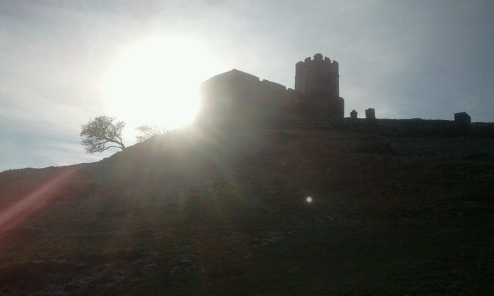 Founded around 1130, the church on Brentor is silhouetted against the sky, with the sun beaming around the east end. A couple of tombstones on the right and a weathered hawthorn tree on the left are also visible as silhouettes