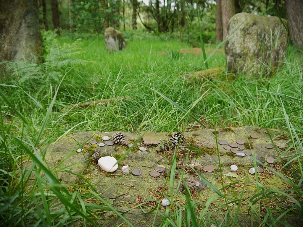 In the foreground, a recumbent stone holds offerings of coins, pebbles, and fir cones. Behind this can be seen some of the weathered, lichen-covered standing stones of a stone circle in a grassy woodland clearing.