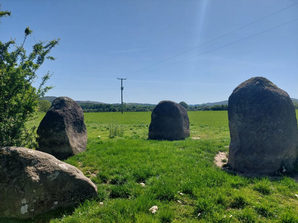 Four small standing stones of dark grey, lichen-covered rock placed in a green, grassy field with hills in the distance