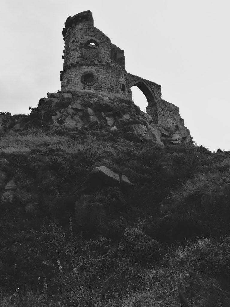 Looking up from the base of a hill, a strange ruin of arches and round tower sit on a rocky outcrop against a dull sky. This is the folly, built in 1752, known as Mow Cop