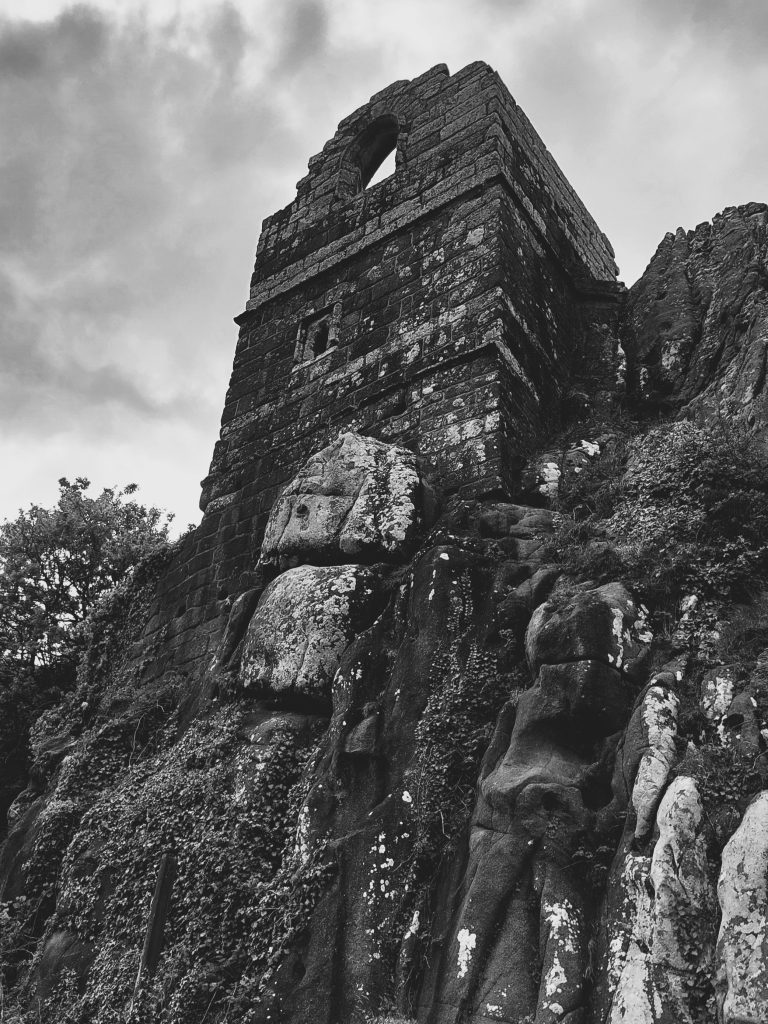 Looking up from the base of a hill, a strange ruin of arched window and square tower sit on a rocky outcrop against a dull sky. This is the remains of a medieval chapel, dating from 1409, at Roche Rock