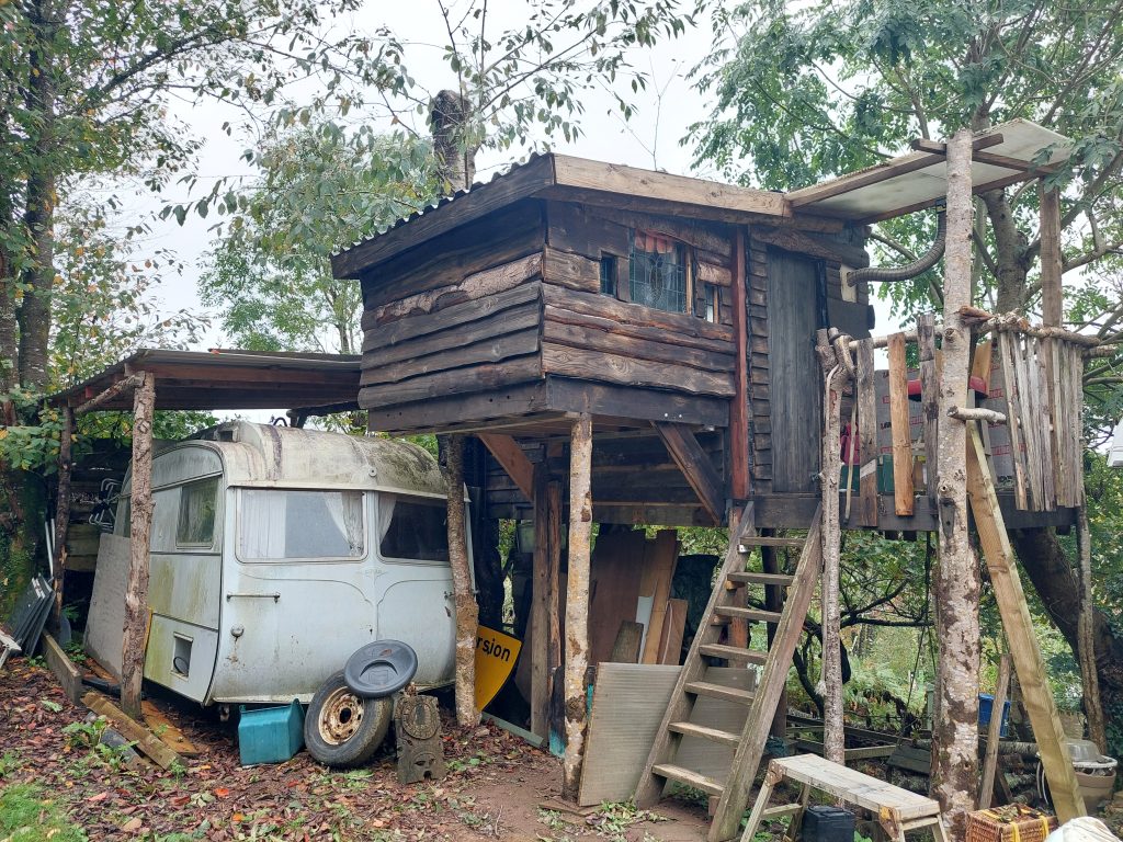 A ramshackle treehouse, made from rough logs and planks, with wooden steps up to a mini veranda outside the door. Beneath the treehouse, under a shelter which forms part of the same structure, sits a vintage Carlight caravan, awaiting renovation